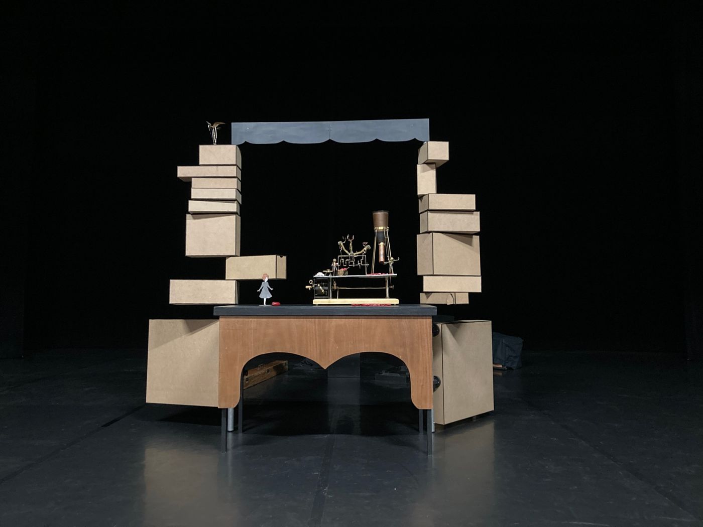 Décor de spectacle de marionnettes : une table en bois et des boîtes en MDF empilées autour d'un cadre noir, sur lequel repose une petite machine de cuivre et de laiton et un personnage découpé